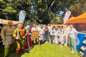 Grupa osób pozuje na trawniku podczas plenerowego wydarzenia. Część osób ma historyczne stroje i rekwizyty, a w tle widać namioty, flagi „Śląsk Cieszyński” oraz stoisko z materiałami promocyjnymi.
