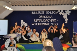 Grupa osób występuje na scenie podczas jubileuszu 10-lecia Ośrodka Rewalidacyjno-Wychowawczego. Część uczestników siedzi lub klęczy i wykonuje gesty rękami, a obok stoją osoby wspierające występ. W tle widoczny jest granatowy napis jubileuszowy oraz białe dekoracje kwiatowe.