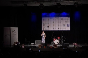 Młoda kobieta śpiewa na oświetlonej scenie podczas wydarzenia artystycznego; w tle widać baner sponsorów, zestaw perkusyjny oraz dekoracje ustawione.