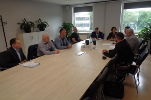 Dziewięć osób w formalnych strojach uczestniczy w spotkaniu przy dużym stole w nowoczesnej sali konferencyjnej. Panuje skupiona atmosfera, na stole woda i notatki, a w tle widać rośliny, okna z widokiem na zieleń i budynki.