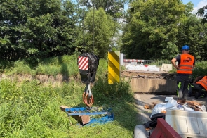 Na drodze widać miejsce prac remontowych przy moście. Obok ustawione są bariery, materiały budowlane oraz element dźwigu z hakiem, a po prawej stronie stoją pracownicy w odzieży ostrzegawczej.