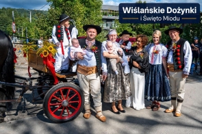 Grupa dorosłych i dzieci pozuje do wspólnego zdjęcia obok udekorowanego wozu zaprzęgowego. Część osób ma na sobie tradycyjne stroje regionalne, a niektórzy trzymają dzieci na rękach. W tle widoczne są drzewa, zabudowania oraz napis „Wiślańskie Dożynki i Święto Miodu”.