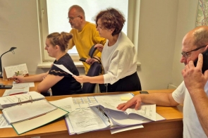  Zdjęcie przedstawia członków sztabu kryzysowego podczas pracy. Przy biurku siedzi Zenon Woźniak, Kierownik  Biura Bezpieczeństwa i Zarządzania Kryzysowego, rozmawia przez telefon. Stoją za biurkiem Starosta Janina Żagan (druga z prawej)  i Wicestarosta Mieczysław Szczurek (drugi z lewej) oraz Marcelina Madzia, podinspektor Biura Bezpieczeństwa i Zarządzania Kryzysowego. Członkowie analizują dokumenty oraz mapę na komputerze. Na biurku widoczne są segregatory, notatki i dokumenty, co podkreśla intensywność pracy i monitorowanie bieżącej sytuacji.