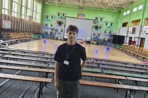 Mężczyzna stoi w centrum hali sportowej. W tle widoczne są ławki.