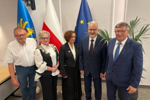Zarząd Powiatu Cieszyńskiego pozuje do zdjęcia w sali przy flagach. W tle widać roślinę oraz fragment stołu.