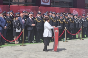 Janina Żagan Starosta Cieszyński w białej marynarce przemawia przy mównicy z logo straży pożarnej. Przed nią stoją równo w szeregu strażacy w galowych mundurach z odznaczeniami, a w tle widnieją czerwone panele z nazwą Komendy Powiatowej PSP.