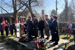 Na niewielkiej scenie stoi grupa osób, w tym mężczyźni w mundurach i garniturach oraz kobieta w płaszczu. Dwóch mężczyzn podaje sobie ręce obok mównicy z herbem gminy. Na scenie leżą wieńce z czerwonych i białych kwiatów, a w tle stoją harcerze, żołnierze i sztandary.