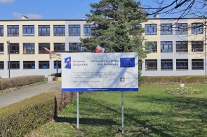 Przed budynkiem szkoły stoi tablica informacyjna z logo Funduszy Europejskich i Unii Europejskiej, zawierająca informacje o dofinansowaniu projektu.