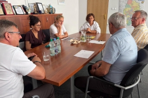 Grupa osób siedzi przy stole. Na stole widać dokumenty, szklanki, dzbanek i butelki z wodą, a w tle szafkę z dyplomami, statuetkami oraz mapę.