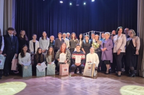 Na scenie pozują uczniowie biorący udział w olimpiadzie razem z Jury oraz współorganizatorzy. Wszyscy są elegancko ubrani. 