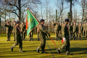 Grupa umundurowanych osób maszeruje na boisku podczas uroczystości. Na pierwszym planie widoczny jest poczet ze sztandarem, a w tle stoją kolejne osoby w szyku.