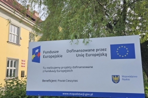 Tablica informacyjna o projekcie dofinansowanym przez Unię Europejską, ustawiona przed budynkiem i drzewami. Na tablicy widoczne są logotypy.