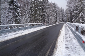 Zaśnieżona droga asfaltowa biegnie przez most otoczony gęstym lasem sosnowym, z barierkami zabezpieczającymi po obu stronach. Świeży śnieg pokrywa drzewa i chodnik, tworząc zimowy krajobraz.