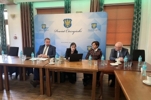 Sala konferencyjna z ustawionymi stołami. Przy stole siedzą cztery osoby, przed nimi znajdują się butelki z wodą, szklanki i laptop. W tle widoczna jest ścianka z herbem oraz napisem „Powiat Cieszyński”.