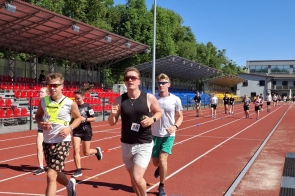 Grupa młodzieży biegnie po czerwonej bieżni lekkoatletycznej na stadionie. W tle widać trybuny z czerwonymi krzesełkami, drzewa oraz słoneczne niebo.