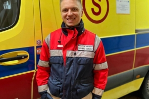 Ratownik medyczny Grzegorz Gabzdyl w czerwono-granatowej kurtce z identyfikatorem „Ratownik Medyczny” stoi przed żółto-czerwoną karetką, uśmiecha się, ma ręce w kieszeniach.