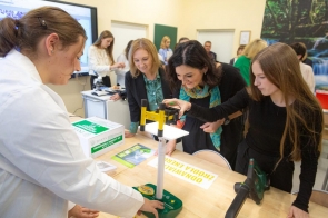 Osoby oglądają wyposażenie pracowni podczas prezentacji. Na stole widoczny jest model turbiny wiatrowej, zestaw edukacyjny dotyczący odnawialnych źródeł energii oraz inne pomoce dydaktyczne.