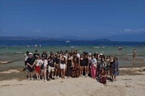 Grupa osób pozuje do zdjęcia na plaży. W tle widać wodę, kąpiących się ludzi, odległy brzeg oraz góry pod błękitnym niebem.