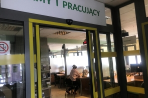 Pomieszczenie z oznaczeniem „FIRMY I PRACUJĄCY”, w którym osoby siedzą przy stanowiskach obsługi. Przestrzeń jest przeszklona, a w środku widoczne są biurka, krzesła i materiały informacyjne.