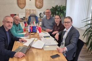 Pięć osób siedzi przy drewnianym stole w biurze. Na stole leżą dokumenty, laptop, telefon i małe flagi państwowe oraz Unii Europejskiej. W tle widoczne są rośliny doniczkowe, okno z żaluzjami oraz elementy wystroju biura.