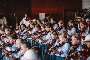 Dzieci i młodzież w strojach regionalnych grają na skrzypcach podczas występu. W tle widać muzyków prowadzących oraz drewnianą scenę.