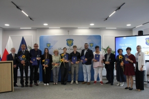 Grupa osób stoi w rzędzie w sali sesyjnej. Część trzyma granatowe teczki z herbem Powiatu Cieszyńskiego, statuetki i słoneczniki. W tle widoczna jest ścianka z herbem powiatu oraz ekran z napisem dotyczącym Gali Sportu.