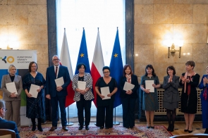 Grupa osób stoi w sali reprezentacyjnej i trzyma dokumenty. W tle widoczne są flagi Polski i Unii Europejskiej oraz baner Kuratorium Oświaty w Katowicach.