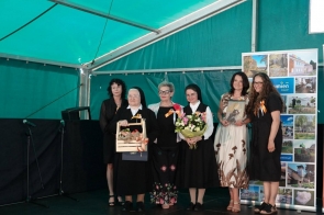 Grupa kobiet pozuje do wspólnego zdjęcia na scenie pod namiotem. Wśród nich są siostry zakonne, jedna z nich trzyma ozdobny upominek, druga bukiet kwiatów, a pozostałe osoby mają przypięte okolicznościowe wstążki.