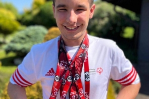 Szymon Machej stoi na zewnątrz w koszulce Olimpiad Specjalnych, uśmiecha się i prezentuje zawieszone na szyi medale zdobyte podczas zawodów.