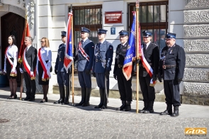 Grupa osób stoi w rzędzie przed budynkiem. Część z nich ubrana jest w mundury strażackie, inne w strój formalny. Kilka osób trzyma sztandary, wszyscy stoją wyprostowani na brukowanej nawierzchni.