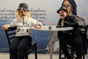 Dwóch młodych chłopców w przebraniach siedzi przy okrągłym stoliku na tle baneru Centrum Edukacyjno-Kulturalnego „U Kossaków” w Górkach Wielkich. Na banerze widać tekst oraz portret kobiety.