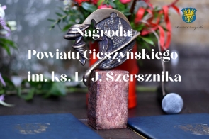 Na pierwszym planie grafiki tekst w kolorze białym – Nagroda Powiatu Cieszyńskiego im. ks. L. J. Szersznika, w tle statuetka wręczana laureatom, składająca się z kamiennego, prostokątnego cokołu oraz mosiężnego odlewu. W prawym górnym rogu herb Powiatu Cieszyńskiego z podpisem.