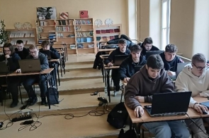 Grupa uczniów siedzi w klasie przy drewnianych ławkach, pracując na laptopach. W tle regały z książkami i dekoracje ścienne. Jasne ściany i duże okna wpuszczają naturalne światło. Niektórzy uczniowie w bluzach z kapturem, skupieni na ekranach komputerów.