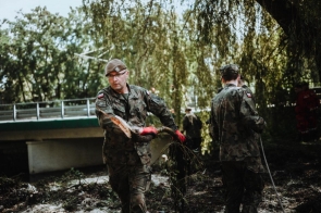 Dwóch żołnierzy w polskich mundurach pracuje w terenie zalesionym. Jeden z nich niesie duży konar z korzeniami, drugi trzyma linę. Obaj mają czerwone rękawice. W tle widoczny jest most, drzewa i inni żołnierze.