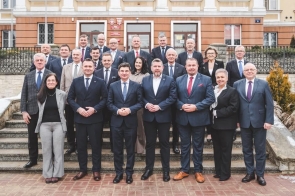 Przed budynkiem na schodach stoi grupa kilkudziesięciu osób ubranych w garnitury i eleganckie stroje. Za nimi widoczna jest elewacja budynku z wejściem i tablicami na ścianie.