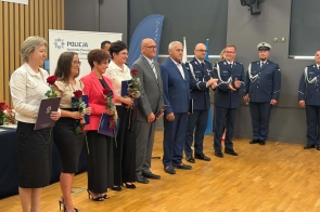 Grupa osób stoi w sali podczas uroczystości. Część osób trzyma róże i teczki, a po prawej stronie stoją policjanci w mundurach. W tle widoczny jest baner Policji oraz godło.