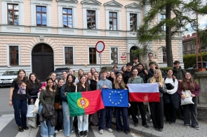 Grupa uczniów stoi przed budynkiem i trzyma flagi Portugalii, Unii Europejskiej oraz Polski, pozując do wspólnego zdjęcia.