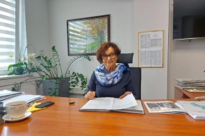 Janina Żagan, Starosta Cieszyński, siedzi przy biurku w gabinecie, wśród dokumentów i kwiatów, z filiżanką herbaty, uśmiechnięta, w okularach i z elegancką apaszką, podpisuje przygotowane pisma.