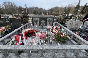 Pomnik z tablicą pamiątkową otoczony wieńcami i zniczami. Wokół zgromadzeni ludzie, część w mundurach. W tle drzewa bez liści oraz zabudowa miejska.