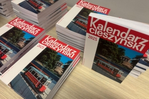 Stosy książek zatytułowanych „Kalendarz Cieszyński 2025” ułożone na jasnym drewnianym blacie. Okładki w odcieniach czerwieni, bieli i błękitu prezentują zdjęcie czerwonego tramwaju na tle budynku otoczonego zielenią. Publikacje są świeżo wydrukowane, równomiernie ułożone w stosy, gotowe do dystrybucji i promocji.
