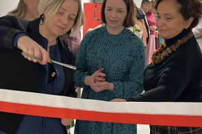 Trzy kobiety stoją przy biało-czerwonej wstędze. Jedna z nich trzyma nożyczki, a w tle widoczne są inne osoby oraz wnętrze budynku.
