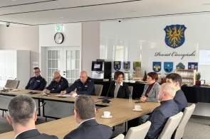 Uczestnicy spotkania siedzą przy stołach ustawionych w podkowę w sali konferencyjnej. Na ścianie widoczny herb, z przodu ekran projekcyjny. Na stołach mikrofony, dokumenty i butelki wody.