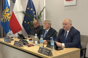 Trzech mężczyzn w garniturach siedzi przy stole konferencyjnym z mikrofonami i tabliczkami. Na stole stoją butelki wody. W tle widoczne flagi Polski i Unii Europejskiej.
