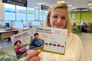 Kobieta trzyma w dłoni materiały zatytułowane „Prawo jazdy małego kierowcy”, ozdobione ilustracjami dzieci i kolorowych samochodów. W tle widoczne są stanowiska obsługi, duże okna oraz tablice z napisami „Rejestracja pojazdów” i „Prawa jazdy”.