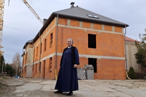 Na tle budowy nowego budynku z czerwonej cegły stoi uśmiechnięta siostra Honorata Klonowska w granatowym habicie z krzyżem na piersi. Po lewej stronie widać żółty dźwig, a wokół placu budowy rozłożone są materiały budowlane. Za budynkiem rosną drzewa iglaste.