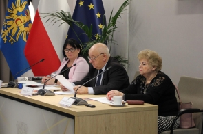 Trzy osoby siedzą przy stole konferencyjnym z mikrofonami; starszy mężczyzna pośrodku czyta dokument, a po jego bokach dwie kobiety uważnie słuchają. W tle znajdują się flagi Polski i Unii Europejskiej oraz roślina.