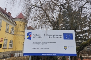 Tablica informacyjna projektu z logotypami instytucji finansujących, ustawiona na zewnątrz, w otoczeniu drzew i zabudowy.