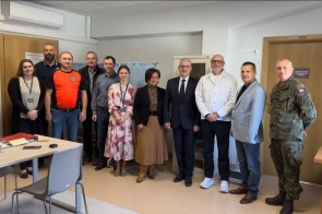 Grupa osób stojących w pomieszczeniu biurowym, ustawionych w rzędzie do wspólnego zdjęcia, wśród nich mężczyzna w mundurze oraz osoby w stroju formalnym i codziennym.