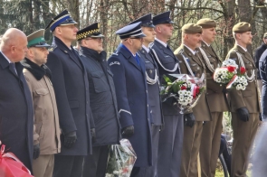 Szereg mężczyzn w mundurach i płaszczach stoi w skupieniu, trzymając wiązanki biało-czerwonych kwiatów podczas uroczystości na świeżym powietrzu.