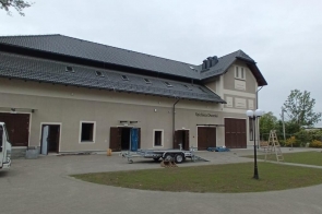 Dwukondygnacyjny budynek z ciemnym, skośnym dachem i jasną elewacją nosi nazwę „Spichlerz Dworski”. Przed nim zaparkowane są auta i przyczepa. Wokół latarnie, trawniki, drzewa i elementy wyposażenia placu.